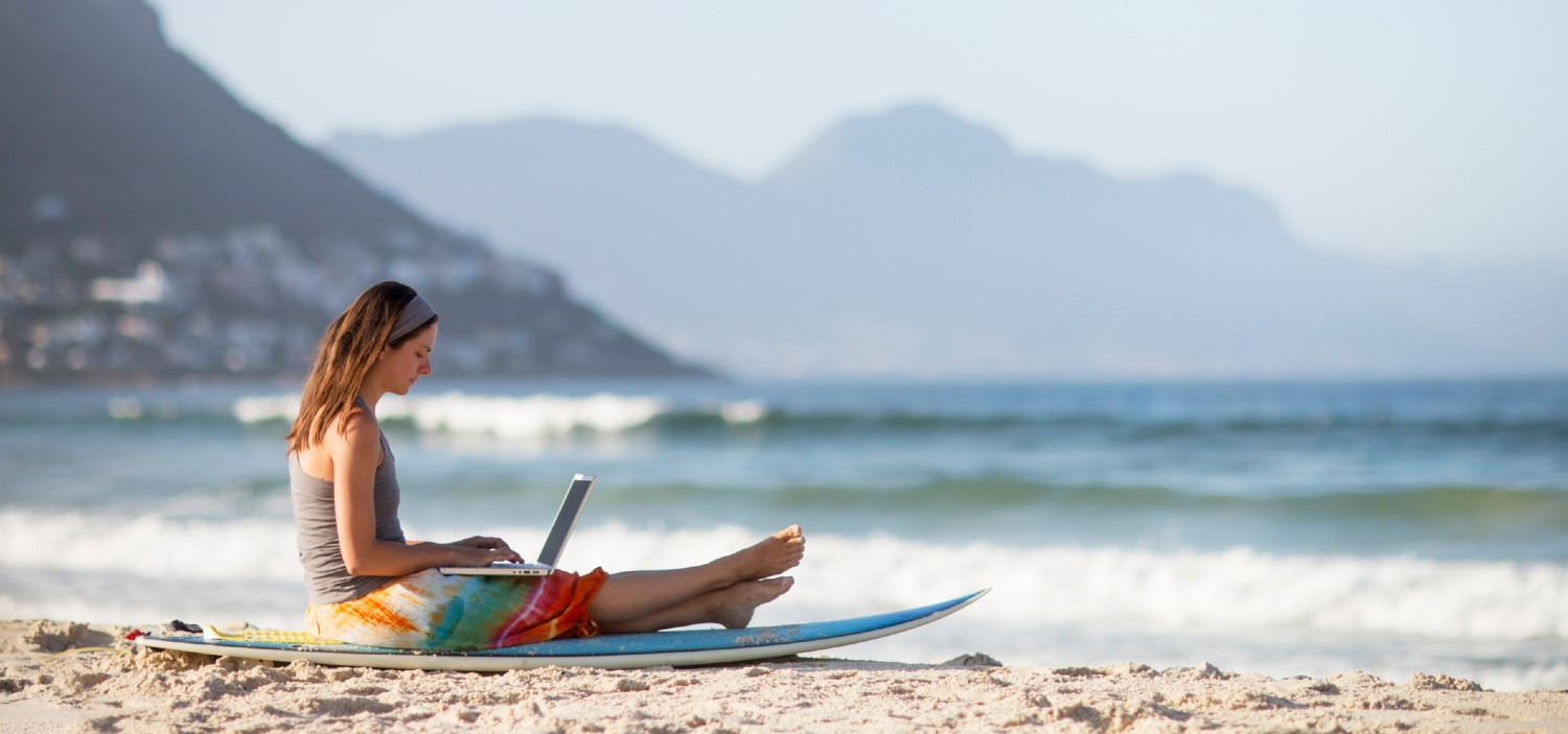 women on laptop sat on surfboard on the beach